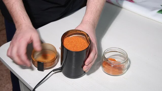 A man pours ground red pepper from a coffee grinder into a jar. Grinding hot pepper. Medium plan.