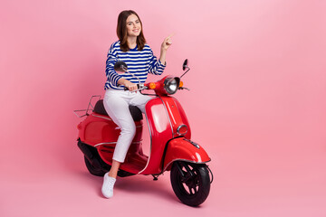 Full body portrait of cheerful nice lady sit scooter indicate finger empty space isolated on pink...