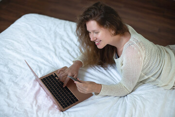 Portrait of happy girl, young positive pretty woman paying, holding credit bank card with phone, laptop. Online shopping at home in living room. Bank application, app.