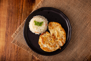 Homemade grilled chicken breast, flattened and seasoned. Accompanied by white rice served in a dark plate on a rustic wooden table. Table top view.