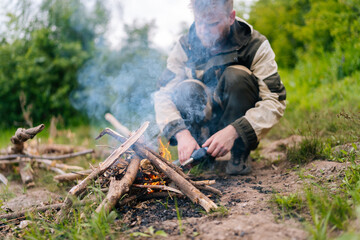 Close-up of blurred survivalist male in raincoat starting fire using gas lighter on bank of river for cooking and warming in evening before sunset. Concept of exploration, travel and adventure. - Powered by Adobe