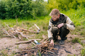 Bearded survivalist male in raincoat putting brushwood on campfire to making fire in forest on cloudy summer day. Concept of scout, research, travel and survival in nature.