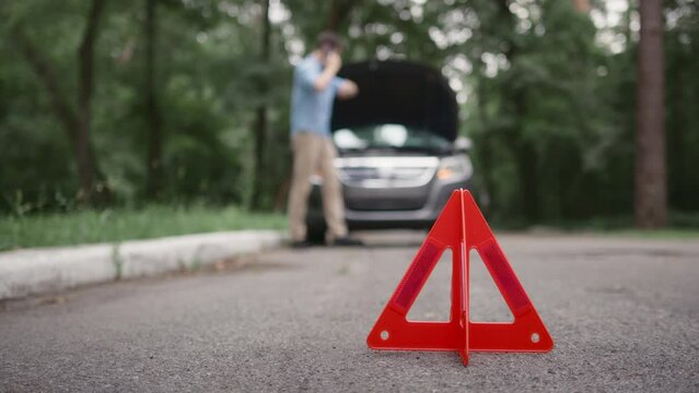 Close up of Red emergency stop triangle sign on road. Upset man calling by smartphone to car service or tow truck for emergency assistance. 