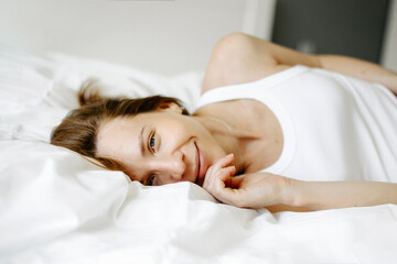 Fototapeta na wymiar Portrait of sensual woman lying on bed in the morning. Attractive girl with natural appearance, dressed in white underwear and looking at camera while relaxing on white blanket