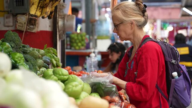 Attractive blonde mature senior woman wearing ethnic attire, buying tomatoes, vegetables and fruit at a local indoor market in Mexico.