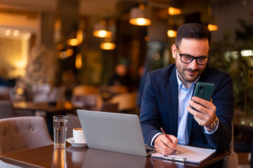 Fototapeta na wymiar Happy smiling businessman wearing suit using laptop and smartphone about to make a deal while sitting and remotely working in modern city restaurant writing notes on clipboard satisfied with the offer