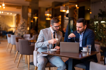 Obraz na płótnie Canvas Businessmen sitting at table in restaurant using laptop, working, thinking of solution. Focused serious male managers analyzing financial report, dedicated to work, improving business strategy.