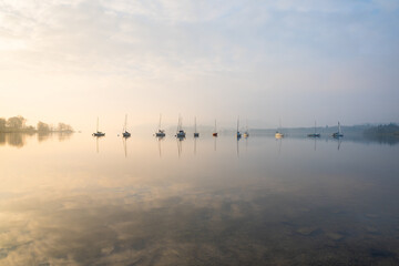 Fototapeta na wymiar Row of yachts sailing boats on a misty morning at Windermere in The Lake District, UK with calm reflections in the water.