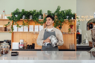 Smiling male bartender wiping a glass and looking away. Barista in a coffee shop holding a towel.