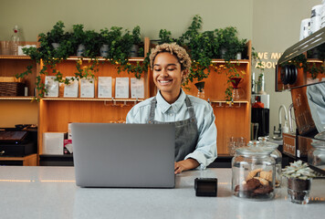 Smiling barista in apron typing on laptop at the counter. Cheerful coffee shop owner working while...