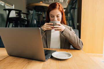 Fototapeta na wymiar Smiling woman with ginger hair enjoying her morning coffee. Businesswoman holding a cup with closed eyes while sitting at a table.