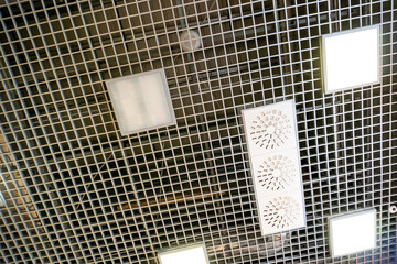 Various lighting fixtures and air conditioning grilles on the ceiling of the restaurant. LED lamps on the suspended mesh ceiling. Industrial air conditioning in the large hall.