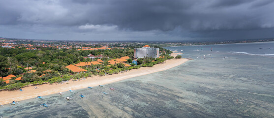Scenic aerial panoramic view at beginning of Sanur beach with wide very shallow shore, dark heavy clouds before rain, Bali, Indonesia