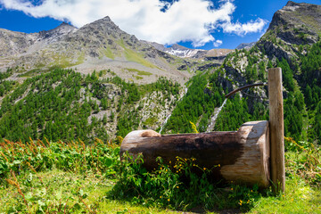 Panorama con primo piano della tipica fontana in legno delle Alpi, e vista su montagne e cascata...