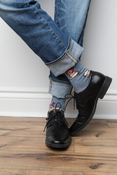 Product photos of men's footwear. Close up photo of a man's foot and legs. He is wearing blue jeans, colorful  blue socks and black dress shoes. His legs are crossed.