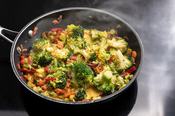 Vegetarian curry with broccoli and other vegetables in a steaming frying pan on a black stovetop,...