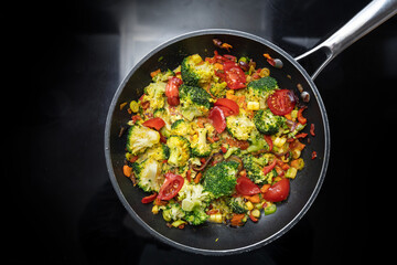 Stir-fried vegetable meal from broccoli, bell pepper, onion and tomato in a frying pan on the black stovetop, vegetarian cooking concept, high angle view from above, copy space