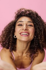 Woman with curly afro hair dancing in pink top and jeans on a pink background, smiling, stretching her arms into the camera, copy space