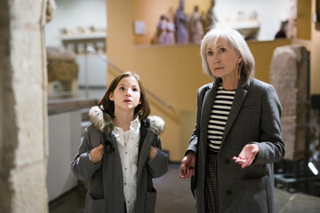 Friendly aged female tutor helping preteen girl exploring antique showpieces in local history museum..