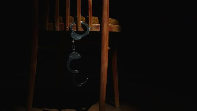 Handcuffs hang and swing chained to chair in dark room.