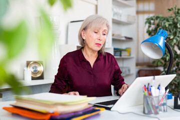 Portrait of busy focused aged female entrepreneur sitting at office desk with papers and laptop ..