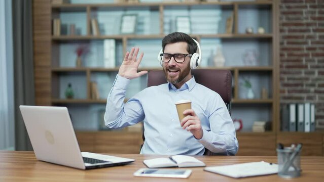 Relaxed mature bearded business man with headphones holding cup of coffee and listening to music and singing at desktop in modern office Happy worker with glasses dancing and having fun indoors alone