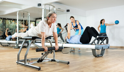 Willing aged woman engaging in pilates training on pedal fitness chair in exercise room during workout session. Persons practicing pilates with trainer