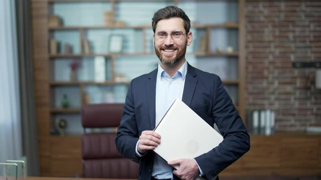 Portrait of confident bearded businessman investor with glasses looking at the camera at modern office workplace Successful smiling entrepreneur owner in formal suit holding laptop computer indoors
