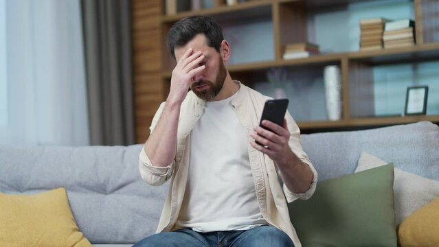 Shocked mature bearded man read bad news while received message email or browsing social media in smartphone at home Worried sad male stares at mobile phone screen with baffled puzzled face indoor 