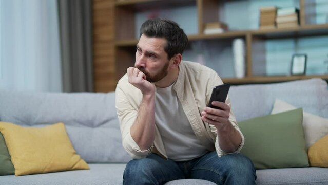 Boring tired bearded man scrolling feed of social networks products in internet store on smartphone sitting on couch in living room Exhausted sad male look at mobile screen without enthusiasm indoor