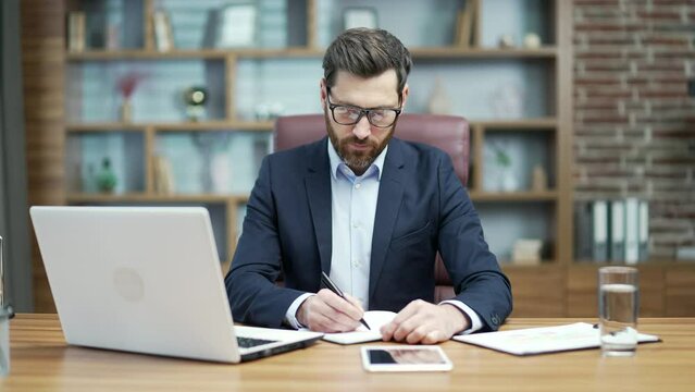 Busy mature business man investor with glasses write notes at organizer planning workday to do list or meeting schedule and typing on laptop computer working online at desktop in modern office indoors