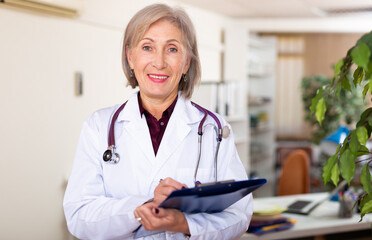 Woman doctor standing in medical office making notes on clipboard