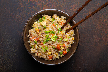 Authentic Chinese and Asian fried rice with egg and vegetables in ceramic brown bowl top view on dark rustic concrete table background. Traditional dish of China