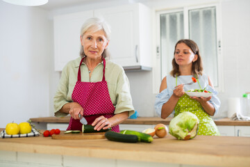 Smiling elderly woman and her daughter preparing fresh vegetable salad at home.