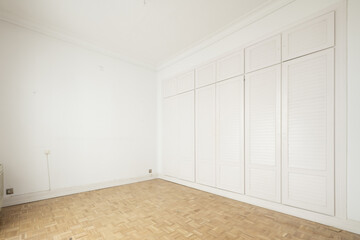 Empty room with one wall covered with white wood cabinets, mezzanine chests all with white Venetian type doors, plain white painted walls and checkerboard shaped oak slatted floors