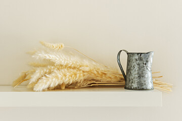 A small old metal jug and dry plants on a white wall shelf