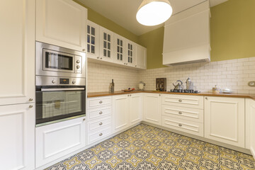 Vintage-style kitchen seen at an angle, furnished in white lacquered wood in the shape of a u with wooden countertop, white stoneware, stainless steel appliances and hydraulic stoneware floor