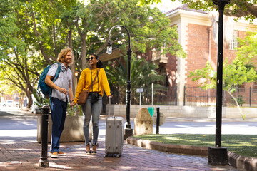 Happy diverse couple with luggage wearing sunglasses and walking in city