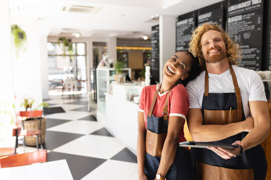 Portrait of happy diverse male and female baristas wearing aprons and using tablet in cafe