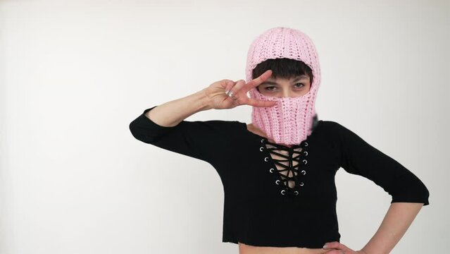 Young woman doing v-sign dance and posing in studio with pink balaclava hat on her head. Fashion industry concept. Copy space. White background. High quality 4k footage