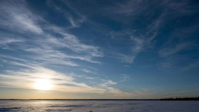 Time lapse of wispy white clouds moving towards the viewer in a blue sky at sunset above a frozen snow covered lake.
