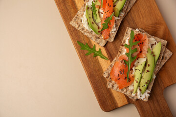 Fresh crunchy crispbreads with cream cheese, salmon, avocado and arugula on beige table, top view. Space for text