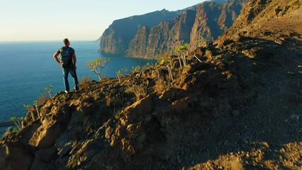 Hiker man with backpack on edge of sheer cliff overlooking ocean. Mountain landscape with seascape. Success well-being achievement concept.