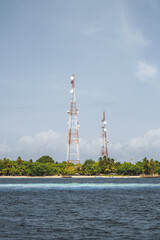 A vertical capture from a yacht overlooking the coast of a private island in the Maldives, on a blue sky day, overgrown with vegetation and two signal repeater towers. In the foreground little beach