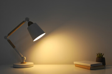 Stylish modern desk lamp, books and plant on table near white wall indoors