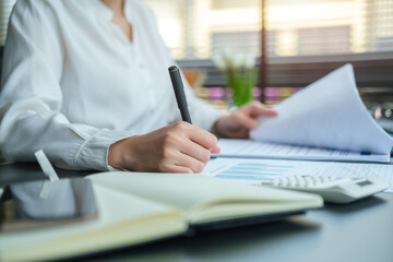 Close up view businesswoman hand holding pen and checking financial reports on office desk..
