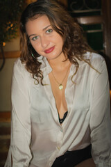 Portrait of a beautiful girl with curly hair in a white dress and black leather shorts sitting on a bar stool. Latina girl with beautiful green eyes