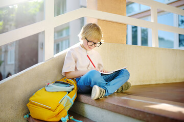 Little student doing homework on break on stair of elementary school building. Portrait of funny nerd schoolboy with big glasses. Vision problems. Back to school concept. Education for kids.