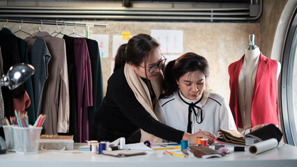 Asian middle-aged female fashion designer teaches a young teen trainee tailor in studio with...