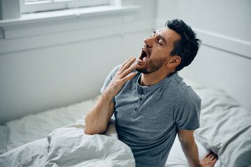 Tired man yawning in bed while waking up in morning at home.